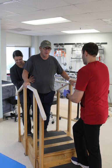 A man wearing a t-shirt, ballcap and dark pants is being helped on a short set of stairs. He's gripping the rails as a woma supports his middle. A man in front of him looks ready to help him if needed.
